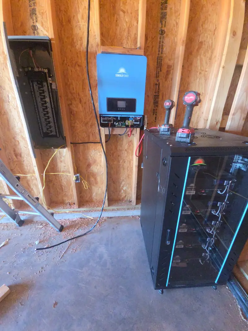 Off-grid power equipment installed in a framed building: a wall-mounted inverter, an open load centre and a rack of batteries.