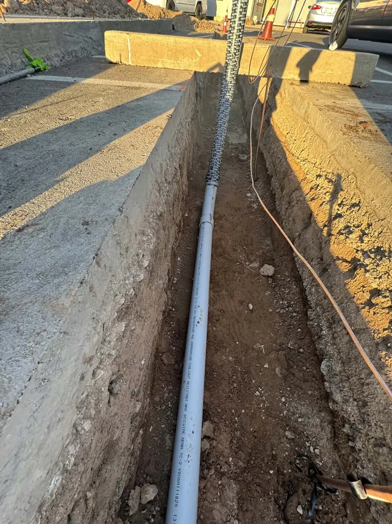 Grey electrical conduit laid along the bottom of an open trench, running toward a riser at the far end.